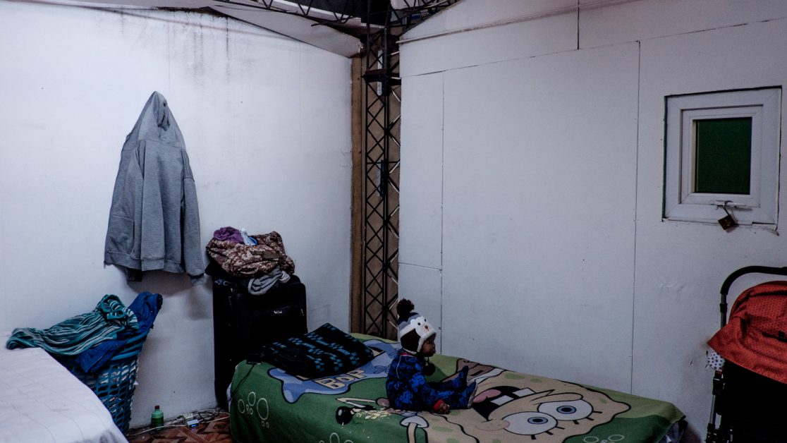 Un niño está sentado en una de las tres camas que hay en su habitación, que mide 12 metros cuadrado y que comparte con otras familias de origen haitiano. Foto: Jorge Vargas | Migrar Photo